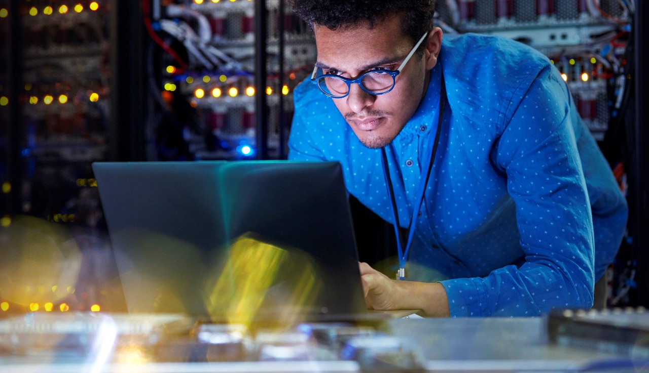 Focused male IT technician working at laptop in dark server room
