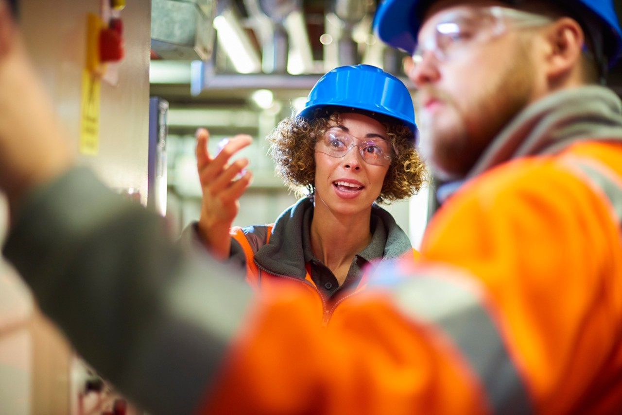 two industrial service engineers conduct a safety check of a control panel and boiler room at a power station . They are both wearing safety equipment and are looking at a control panel . The female is holding a digital tablet and discussing possible solutions to fix the problem with her male colleague.