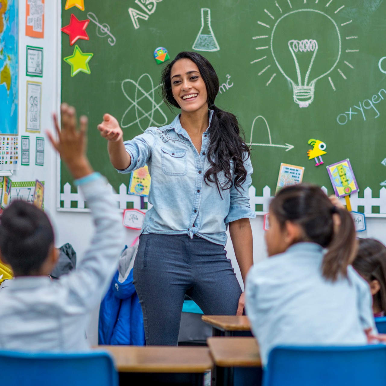 An Ethnic teacher is leading a class of elementary school children. There are various posters on the wall, and drawings on the chalkboard. Students are putting up their hands to answer a question.